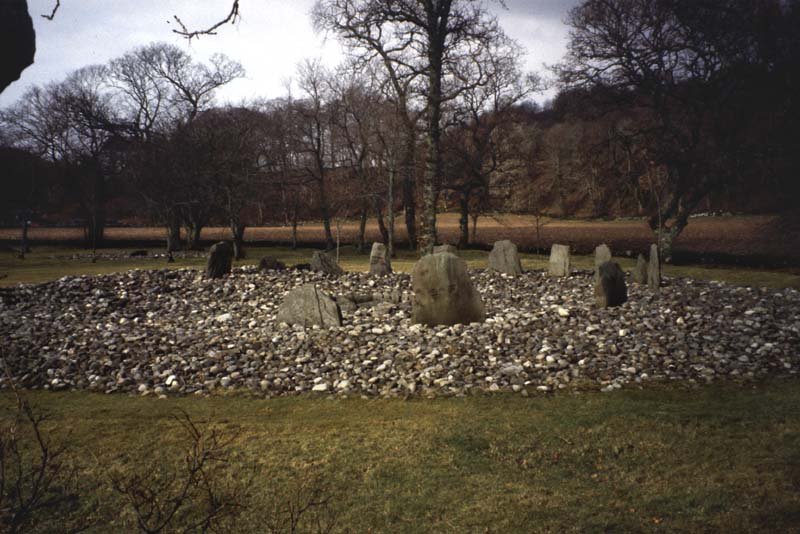 A photograph of a stone circle within the basal remains of a large stone cairn in a grassy landscape with trees in the background